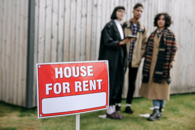 a red "house for rent" sign on a green lawn with prospects standing in the background
